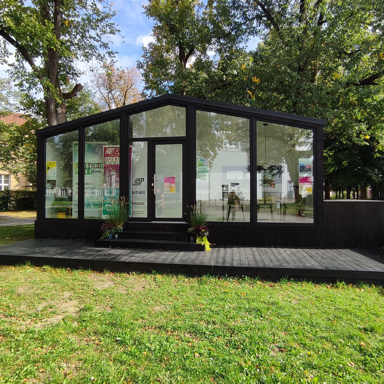 Glasmodernes Tinyhaus mit schwarzen Holzrahmen auf Rasenfläche und herbstlichen Bäumen.