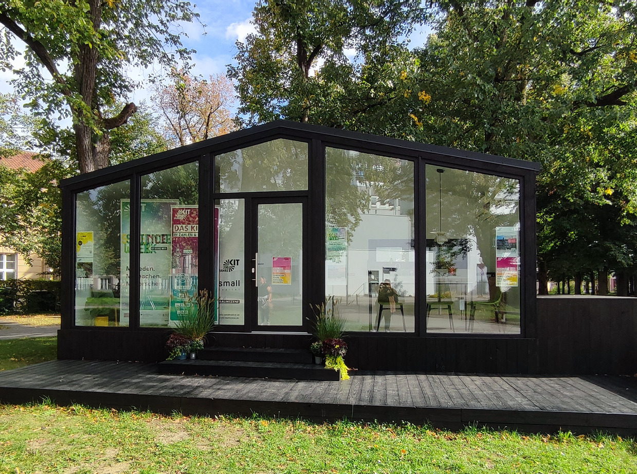 Reiß, KIT Glasmodernes Tinyhaus mit schwarzen Holzrahmen auf Rasenfläche und herbstlichen Bäumen.