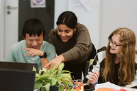Drei junge Menschen lachen und arbeiten gemeinsam an einem Tisch mit Laptops.