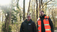 Zwei Männer in Waldumgebung, einer in dunkler Jacke, der andere in orangener Warnweste; Sonnenlicht im Hintergrund.