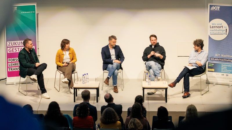 Fünf diskutierende Personen sitzen auf einer Bühne in einem Panel-Format, Zuschauer davor.